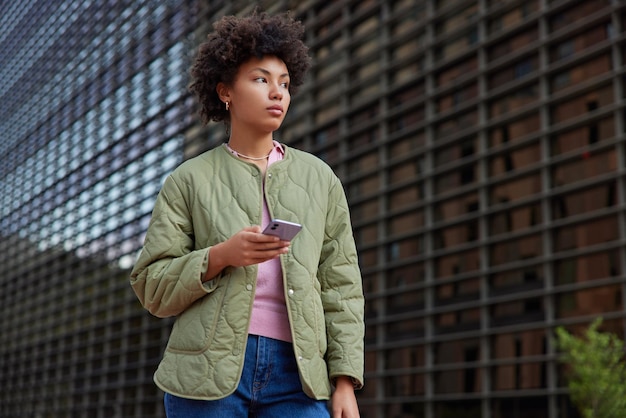 Foto de mujer guapa con chaqueta jeans sostiene teléfono móvil instala aplicación actualiza perfil en redes sociales mira hacia otro lado pensativamente al aire libre