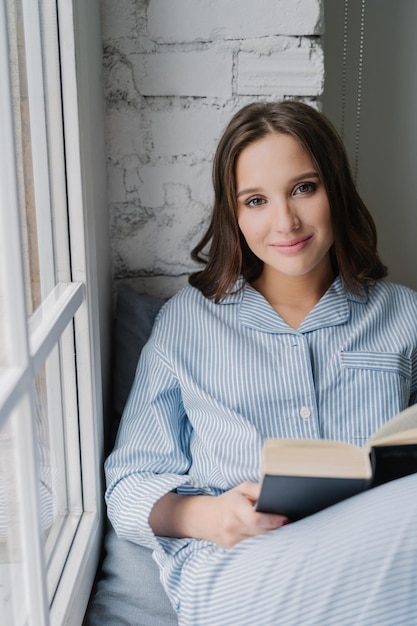 La foto de una mujer hermosa vestida con ropa informal usa pijamas se sienta en el alféizar de la ventana lee una historia romántica en un libro disfruta de la comodidad y la comodidad Mujer joven con libro de texto en casa Concepto de ocio