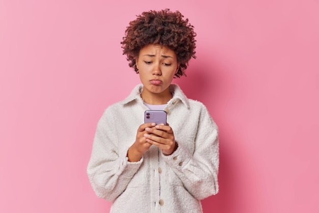 La foto de una mujer infeliz de pelo rizado usa un aparato moderno y se ve decepcionada con la pantalla del teléfono inteligente.