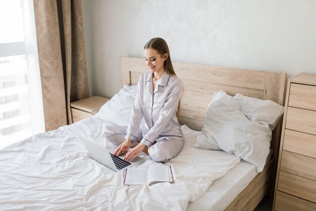 Foto de mujer joven alegre en pijama escribiendo en la computadora portátil y sonriendo mientras está sentado en la cama en la habitación