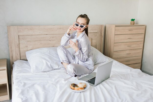 Foto de mujer joven alegre en pijama con laptop y sonriendo mientras está sentado en la cama en la habitación de luz