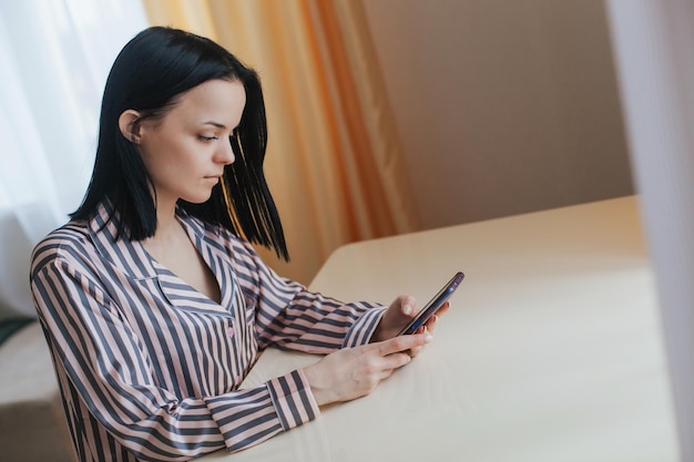 Foto de una mujer joven y atractiva en pijama sentada en el interior de una mesa en un ambiente hogareño agradable por la mañana con un teléfono móvil en las manos