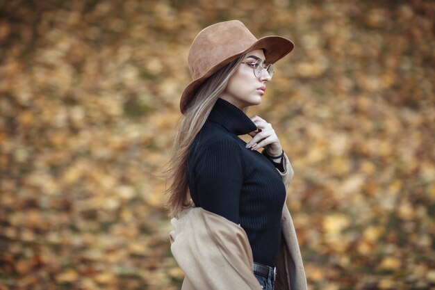 Foto de una mujer joven y atractiva vestida con un abrigo y sombrero de fieltro en el fondo de hojas caídas en el parque de otoño