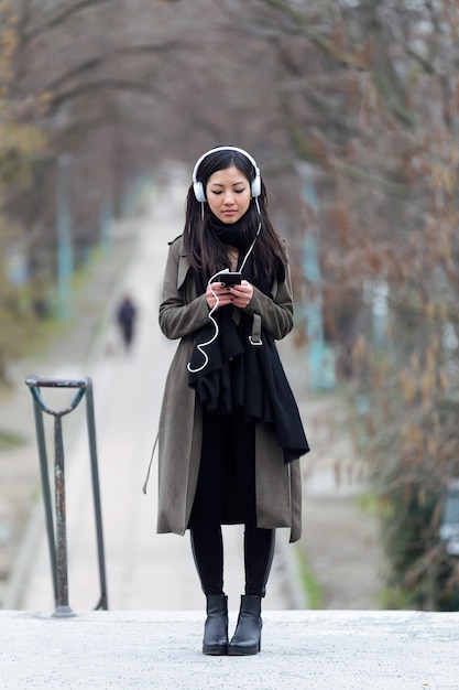 Foto de mujer joven bastante asiática escuchando música con su teléfono inteligente en la calle.