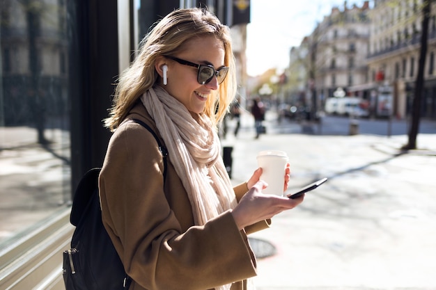 Foto de mujer joven y bonita escuchando música con auriculares inalámbricos y el teléfono inteligente en la calle.