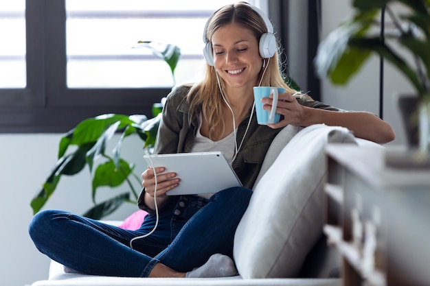 Foto de mujer joven y bonita escuchando música con su tableta digital mientras bebe una taza de café en el sofá en casa.