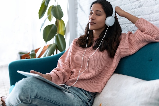 Foto foto de mujer joven y bonita escuchando música con tableta digital y relajarse mientras está sentado en el sofá en casa.