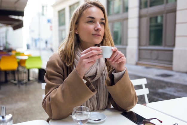 Foto de mujer joven y bonita mirando hacia los lados mientras bebe una taza de café en la terraza de una cafetería.