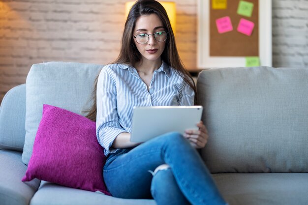 Foto de mujer joven y bonita que usa su tableta digital para leer mientras está sentado en el sofá en casa.