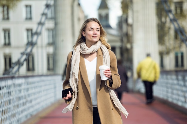 Foto de mujer joven y bonita sosteniendo una taza de café y un teléfono inteligente mientras caminaba por la calle.