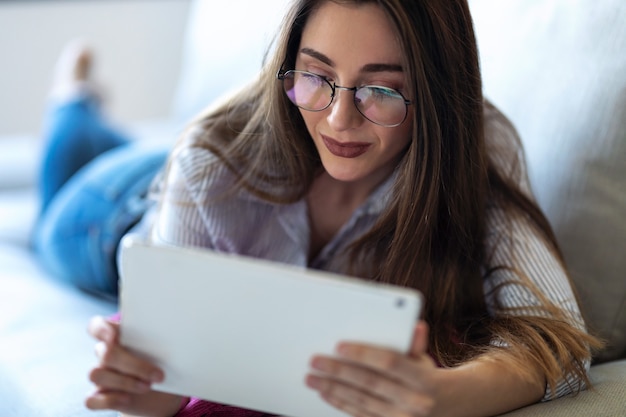 Foto de mujer joven y bonita con su tableta digital para leer mientras está acostado en el sofá en casa.