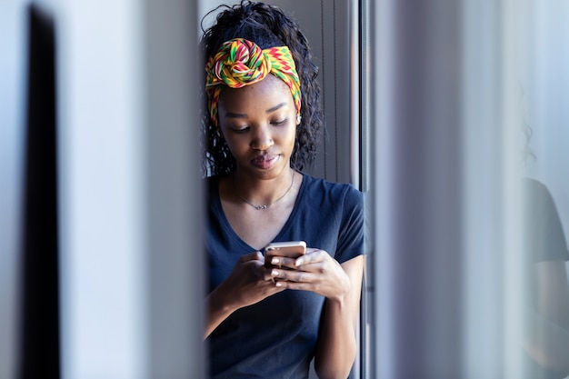 Foto de mujer joven y bonita con su teléfono móvil mientras está de pie junto a la ventana en casa.