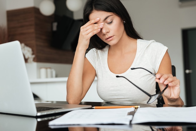 Foto de mujer joven cansada analizando las finanzas del hogar con un portátil mientras sufre fatiga visual y sostiene gafas.