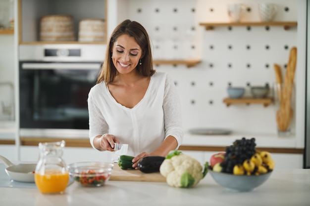 Foto de una mujer joven cortando verduras y preparando una comida saludable en su cocina.