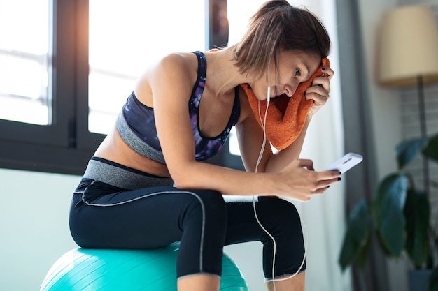 Foto de mujer joven deportiva con su teléfono móvil después de la sesión de ejercicios mientras está sentado en la pelota de fitness en casa.