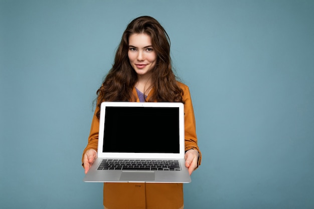 Foto de mujer joven feliz seria hermosa que sostiene el ordenador portátil que mira la cámara que lleva la chaqueta amarilla aislada sobre fondo azul de la pared. copia espacio