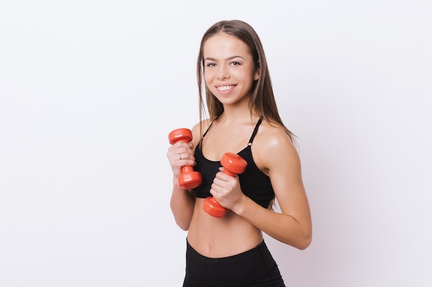 Foto de mujer joven en forma sosteniendo pesas rojas, mientras mira a la cámara con una sonrisa dentuda, de pie sobre fondo blanco.