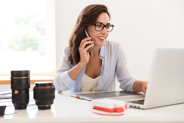 Foto foto de una mujer joven fotógrafa optimista alegre en la oficina trabajando por computadora portátil y tableta gráfica hablando por teléfono móvil.