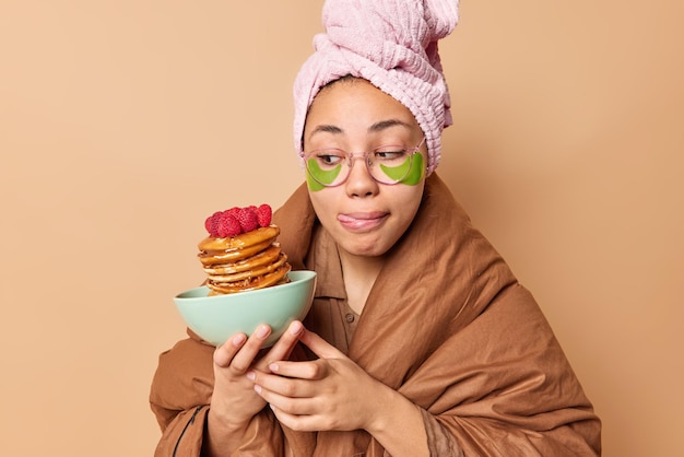 La foto de una mujer joven se lame los labios usa una toalla envuelta y una manta mira el tazón de panqueques con frambuesas que van a desayunar aplica parches de hidrogel verde aislados sobre fondo marrón