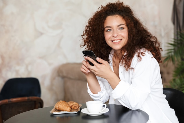 Foto de una mujer joven optimista feliz vestida con camisa blanca en el interior en el hotel en casa en la cocina tomando café comer croissant desayunar con teléfono móvil.