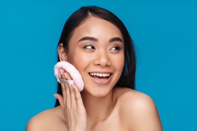 Foto de mujer joven optimista positiva asiática posando aislada en la pared azul con borla de polvo.