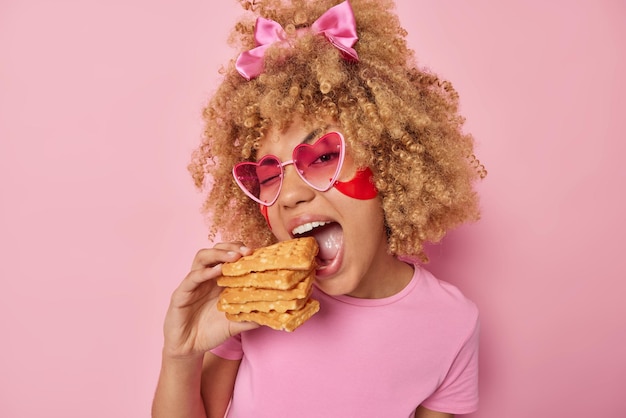 Foto foto de mujer joven de pelo rizado muerde gofres caseros guiños ojo usa gafas de sol en forma de corazón y camiseta casual aislado sobre fondo rosa bonita modelo femenina disfruta de postre favorito
