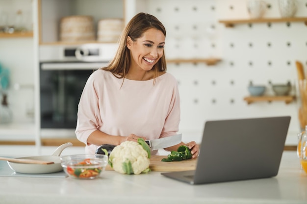 Foto de una mujer joven que usa una computadora portátil mientras prepara una comida saludable en casa.