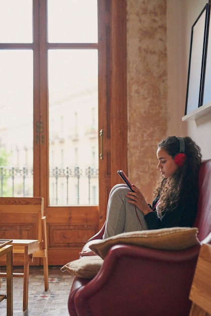 Una foto de una mujer joven que usa una tableta digital y auriculares en el sofá de casa