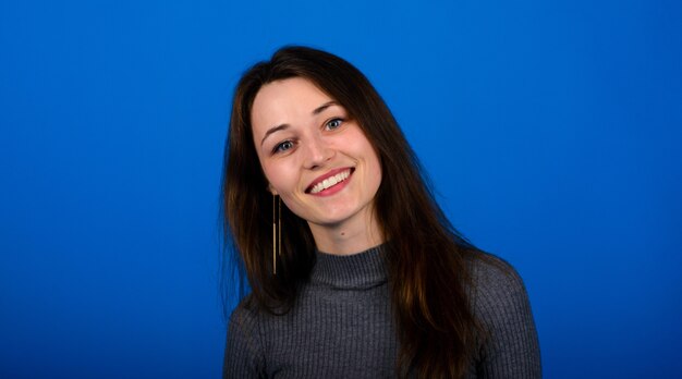 Foto de mujer joven sonriente y alegre en vestido gris sobre fondo azul. Retrato emocional