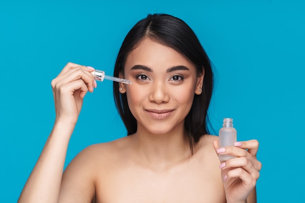 Foto de mujer joven sonriente asiática posando aislada en la pared azul con suero amplio para cuidar su piel.