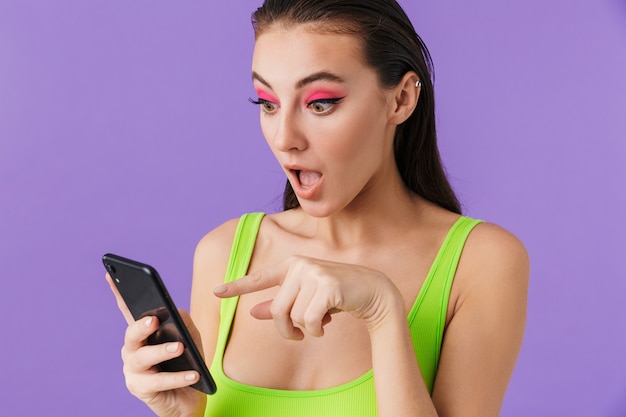 Foto de mujer joven sorprendida con maquillaje brillante usando y apuntando con el dedo en el teléfono inteligente