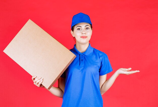 Foto de mujer joven en traje azul con caja de cartón sobre pared roja.