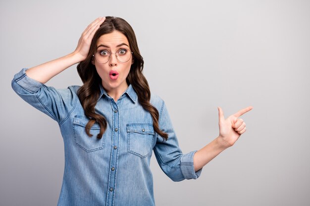 Foto de mujer loca de negocios con la boca abierta indican el espacio vacío del dedo aconsejando precios bajos con el brazo en la cabeza, especificaciones de desgaste, jeans casuales, camisa de mezclilla, color gris aislado