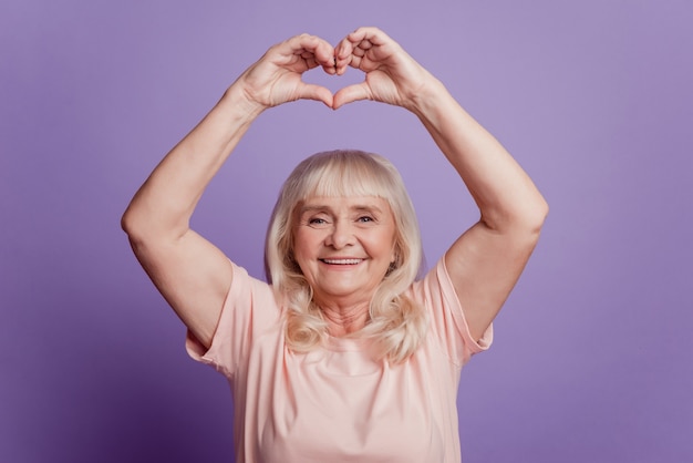 Foto de mujer madura demuestran los dedos del corazón firman por encima de la cabeza sobre fondo violeta