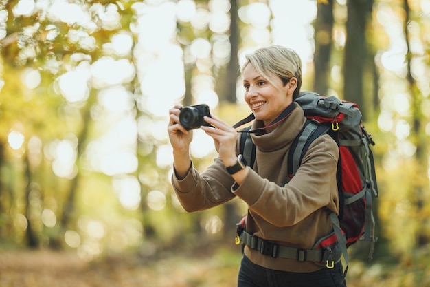 Una foto de una mujer madura tomando una foto de un entorno con una cámara digital durante un paseo por el bosque en otoño.