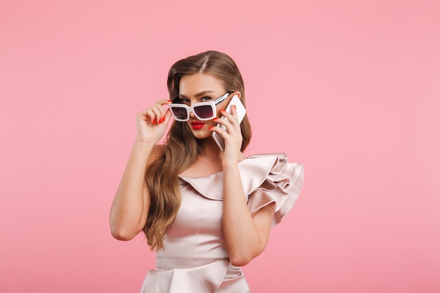 Foto de mujer de moda sensual de 20 años en vestido hablando por teléfono inteligente y tocando gafas de sol de moda, aisladas sobre pared rosa