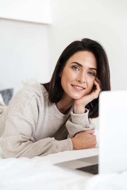 Foto de mujer morena de 30 años usando laptop, mientras está acostado en la cama con ropa blanca en una habitación luminosa