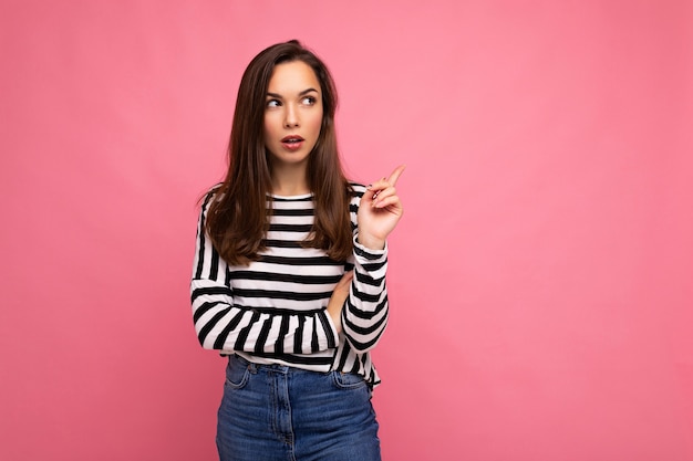 Foto de mujer morena atractiva pensativa feliz positiva joven con emociones sinceras vistiendo manga larga a rayas casual aislado sobre fondo rosa con espacio vacío y pensamiento.