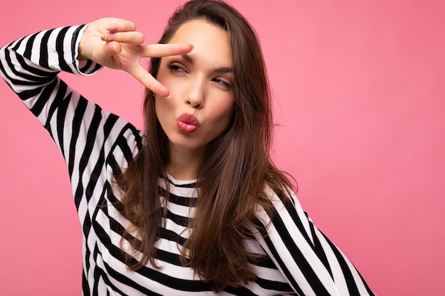 Foto de mujer morena hermosa feliz encantadora positiva europea joven con emociones sinceras vistiendo jersey de rayas casual aislado sobre fondo rosa con espacio de copia y dando beso.