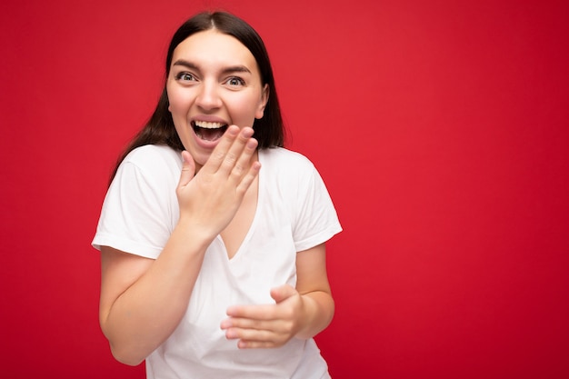 Foto de mujer morena hermosa joven positiva con emociones sinceras vistiendo camiseta blanca para maqueta aislada sobre fondo rojo con espacio vacío y riendo