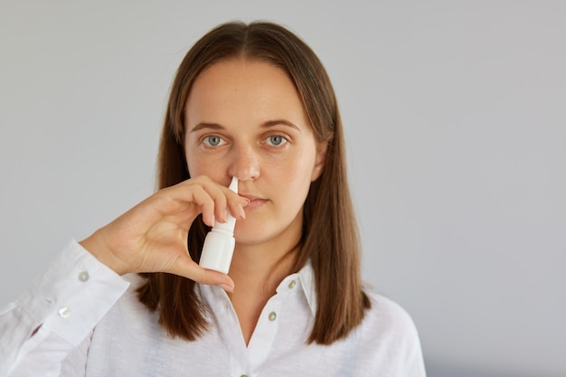 Foto de mujer morena insatisfecha, usa spray nasal, sufre de congestión nasal, vestida de blanco, posando en interiores contra una pared clara, mirando a la cámara.