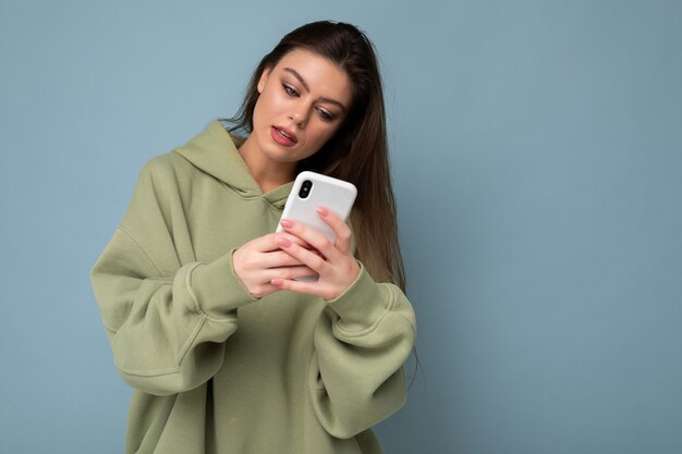 Foto de mujer morena joven hermosa seria vistiendo elegante sudadera con capucha verde mediante teléfono móvil