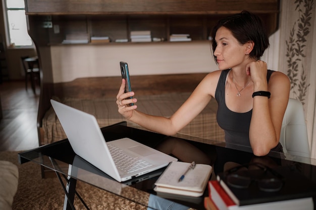 Foto una foto de una mujer muy joven que usa su teléfono móvil mientras trabaja con una laptop sentada en casa