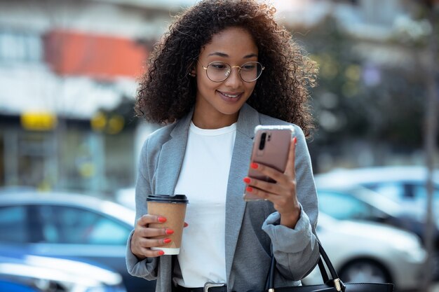 Foto de mujer de negocios bastante joven con su teléfono inteligente mientras toma café caminando por la calle.