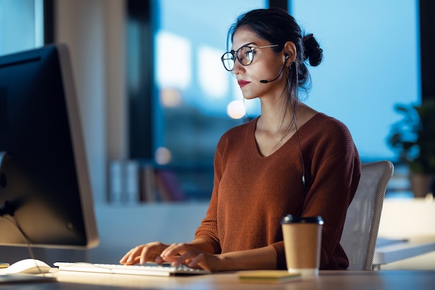 Foto de mujer de negocios joven hermosa que trabaja con la computadora mientras habla con el auricular sentado en la oficina.