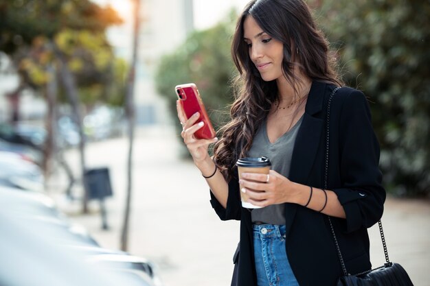 Foto de mujer de negocios joven hermosa usando su teléfono móvil mientras toma café caminando por la calle.