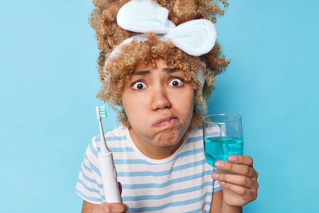 La foto de una mujer preocupada y sorprendida sostiene un cepillo de dientes eléctrico y un vaso de enjuague bucal fresco usa una diadema y una camiseta casual a rayas se somete a rutinas diarias de higiene bucal aisladas sobre una pared azul