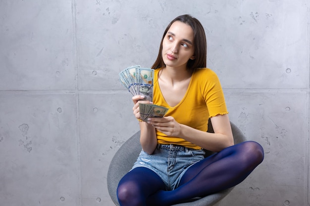 Foto de una mujer rica con ropa sencilla sosteniendo un abanico de dinero en dólares aislado contra un fondo de pared de hormigón