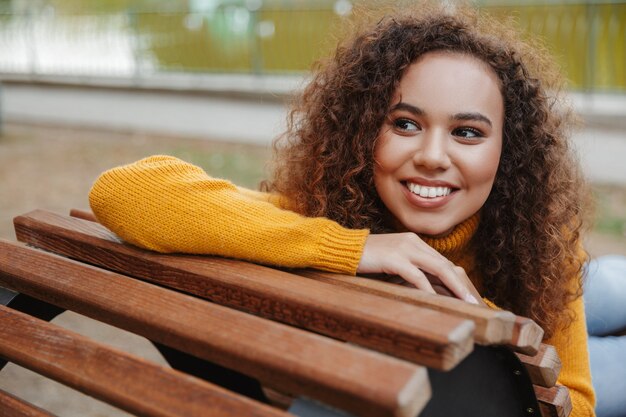 Foto de una mujer rizada joven hermosa alegre feliz positiva sentarse en un banco en el parque al aire libre.