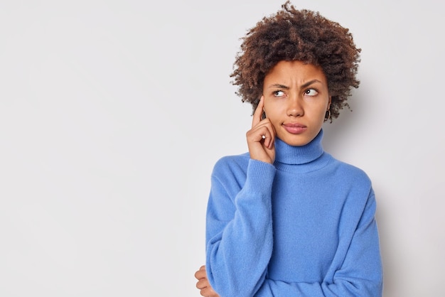 Foto de mujer rizada joven seria mantiene el dedo en la sien intenta concentrarse considera que algo contempla sobre algo importante viste un puente azul casual aislado sobre fondo blanco.